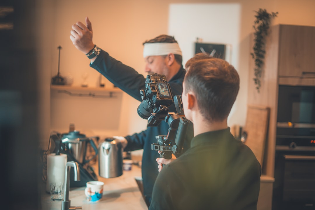 Man with camera filming another man in kitchen.