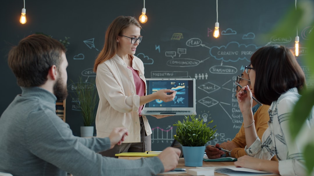 Girl office worker is making presentation pointing at laptop screen talking to young people colleagues. Modern technolog