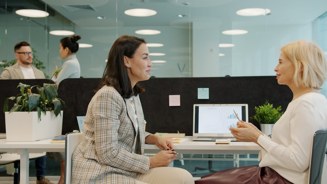 Cheerful women coworkers talking discussing work looking at laptop screen sitting at desk in open space office room. People a